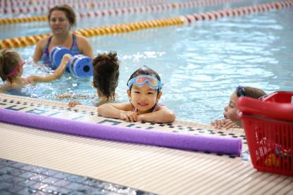 a small child with their arms up on the side of the pool. There is a purple pool noodle in the foreground
