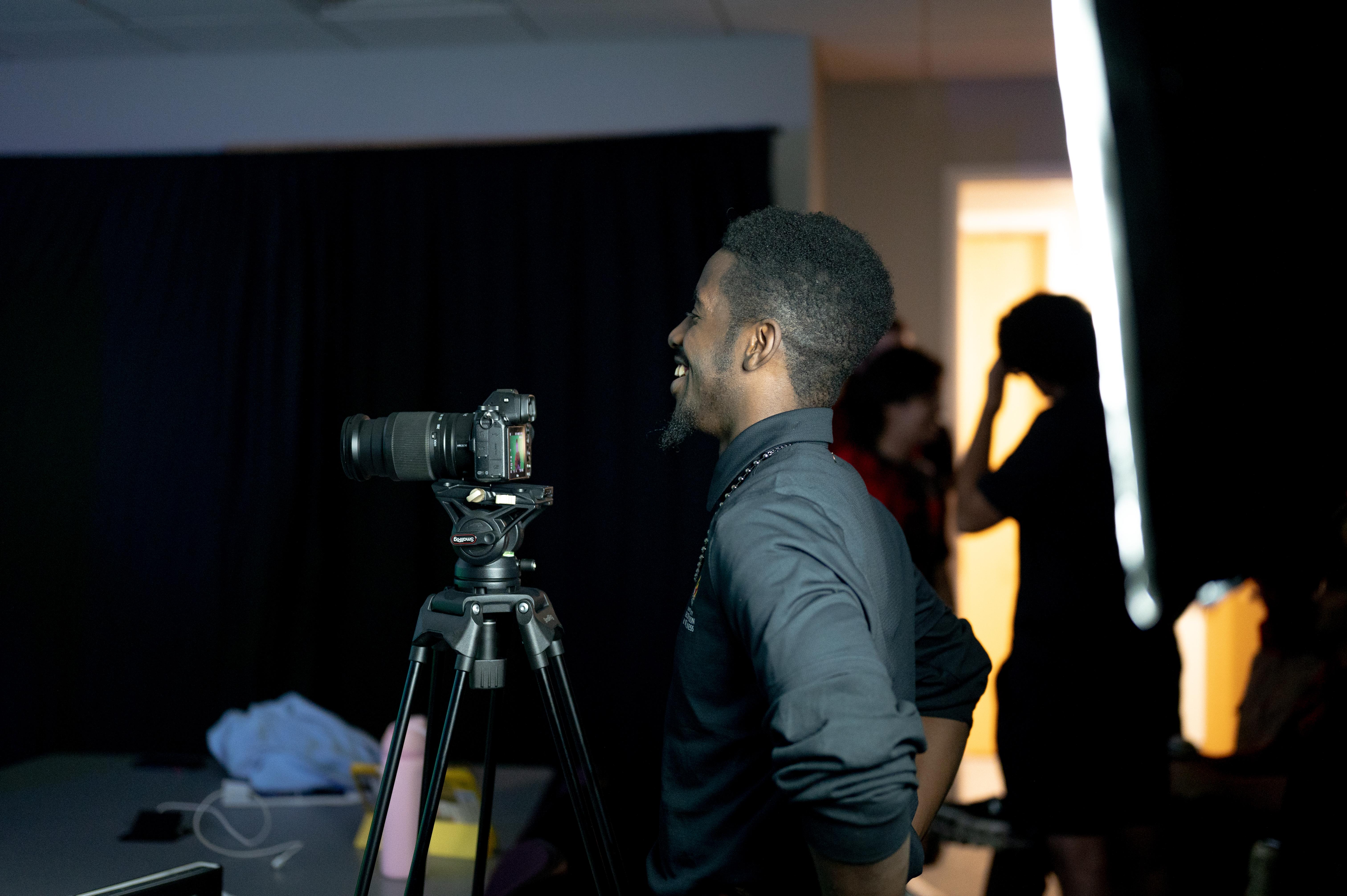 Male content team staff member, smiling at the subjects in front of the camera who are out of frame, as he stands behind the camera with his hands on his hips.